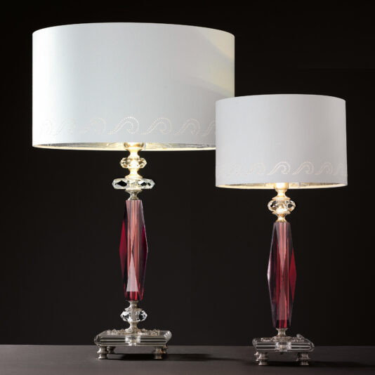 Two sophisticated Luxury Antique Rose Glass Table Lamps, adorned with white drum lampshades and featuring crystalline bodies with a striking red, elongated central segment, are displayed against a dark backdrop. Their meticulous yet contemporary design is emphasized by the light they emit. Notably, one lamp stands taller than the other.