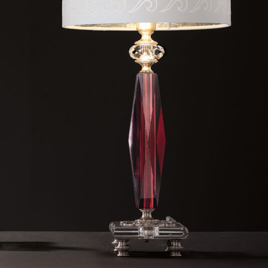 The Luxury Antique Rose Glass Table Lamp, featuring a decorative crystal base and a red glass cylindrical column, glows against a dark background. Its light-colored lampshade with subtle patterns and intricately detailed metal base create an exquisite blend of vintage elegance.