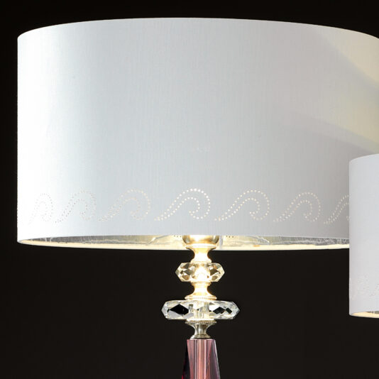 A close-up view of the Luxury Antique Rose Glass Table Lamp reveals a white lampshade with delicate perforated patterns near the bottom edge, a clear crystal base with intricate detailing, and a metallic central rod. The dark background accentuates the illuminated antique rose glass table lamp.