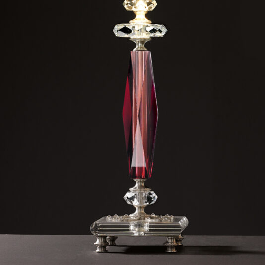 The Luxury Antique Rose Glass Table Lamp features an elegant, slender, faceted base in deep red glass adorned with crystal accents and metallic details. It stands gracefully on a small ornate platform with four feet. The lit lamp casts a warm glow against the dark background, highlighting its luxurious design.