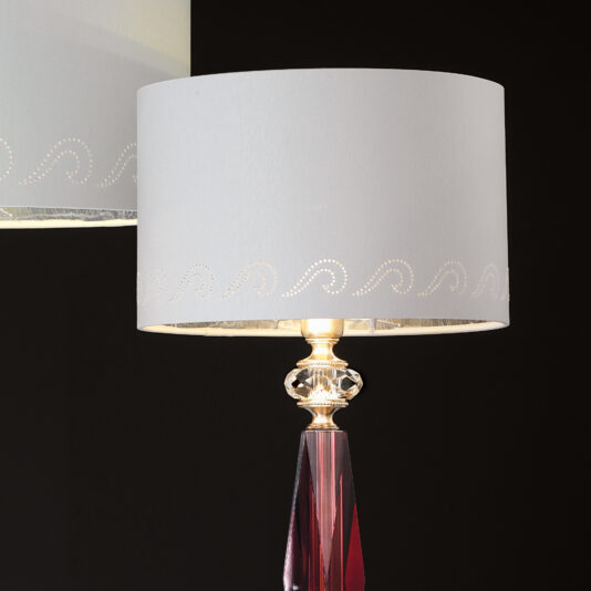 The Luxury Antique Rose Glass Table Lamp boasts a modern cylindrical white lampshade adorned with a subtle wave pattern near the edge and topped with a transparent glass finial, all set against a dark background. Its antique rose-tinted glass base adds an elegant touch to the overall design.