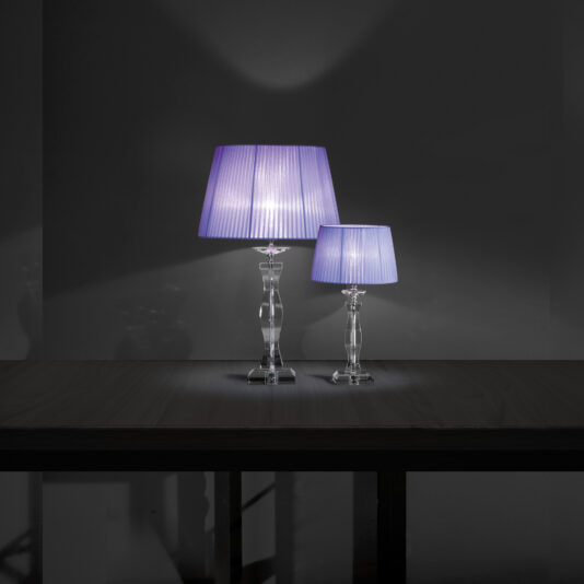 Two Modern Glass Table Lamps With Pleated Shades, one larger and one smaller, featuring elegant clear bases and purple pleated shades, are placed on a dark wooden table. Both lamps are lit, casting a soft glow against a dark grey background.