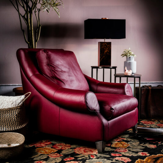 In a cozy room, a Modern Leather Designer Armchair with sleek lines and plush cushions in deep red takes center stage. The backdrop features a black lamp on a dark side table, accompanied by a potted plant. A patterned rug with floral designs adds warmth to the space, complemented by wicker baskets placed beside the chair.
