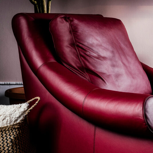 Close-up of the Modern Leather Designer Armchair, showcasing its plush, red leather with smooth curves and intricate stitching details. Beside the chair, a woven basket filled with a white blanket is partially visible. In the background, neutral-colored walls and plant stems reaching from behind set a serene scene.