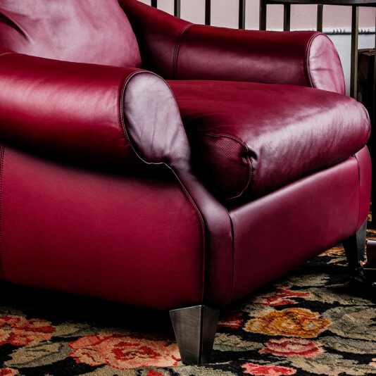 A detailed view of the Modern Leather Designer Armchair in a rich red hue, featuring sleek dark metal legs. The armchair rests on a lively floral-patterned rug, with a railing visible in the background. The smooth, well-cushioned leather exudes both comfort and sophistication.
