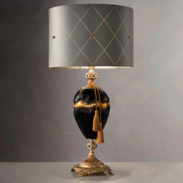 Traditional Amber And Blue Glass Table Lamp Introducing the Traditional Amber And Blue Glass Table Lamp, a luxurious piece featuring an amber and blue glass vase-shaped base adorned with gold accents and a golden tassel. It stands on an ornate, brass-colored pedestal and is topped with a gray lampshade designed with a diamond pattern and small golden accents. The background showcases a plain gray wall to complement its elegance.