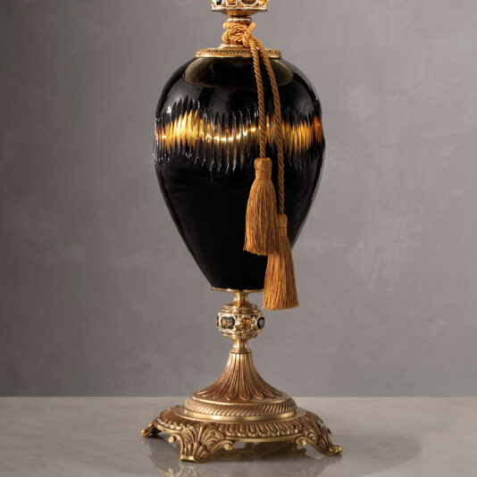 A vintage black ceramic vase with a glossy finish, adorned with a golden tassel, stands on an ornate gold pedestal featuring intricate detailing. This setup exudes the same traditional elegance as the Traditional Amber And Blue Glass Table Lamp set against a plain gray backdrop.