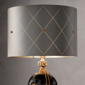 Traditional Amber And Blue Glass Table Lamp The Traditional Amber And Blue Glass Table Lamp boasts a patterned grey lampshade adorned with diamond shapes and subtle golden highlights. Its base, painted in black with gold accents and detailed craftsmanship, features an elegant gold rope around the neck. The lamp is set against a plain grey backdrop.