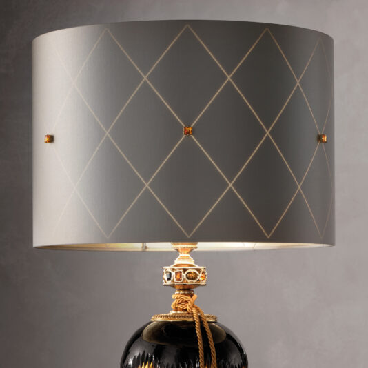 The Traditional Amber And Blue Glass Table Lamp boasts a patterned grey lampshade adorned with diamond shapes and subtle golden highlights. Its base, painted in black with gold accents and detailed craftsmanship, features an elegant gold rope around the neck. The lamp is set against a plain grey backdrop.