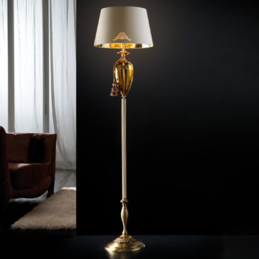 Traditional Amber Blown Glass Floor Lamp A luxurious floor lamp with a gold base stands in a dimly lit room. The **Traditional Amber Blown Glass Floor Lamp** features an elegant white shade with gold accents and a decorative tassel. In the background, part of a brown upholstered chair and a window with sheer white curtains are visible.