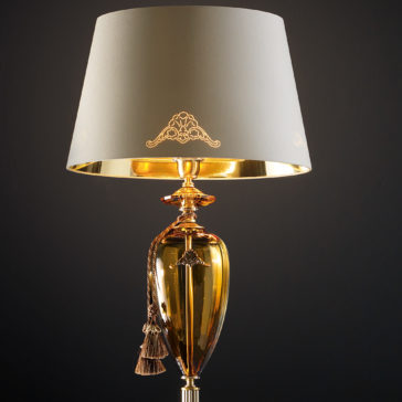 Traditional Amber Blown Glass Floor Lamp A detailed photograph of the Traditional Amber Blown Glass Floor Lamp showcasing an ornate amber-tinted blown glass base and a cream-colored lampshade. The lampshade is adorned with intricate gold designs, and a gold tassel embellishes the top of the base. The background is solid black.