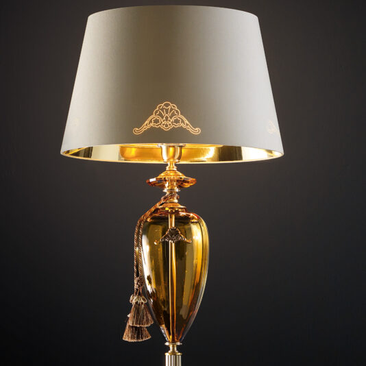 Traditional Amber Blown Glass Floor Lamp A detailed photograph of the Traditional Amber Blown Glass Floor Lamp showcasing an ornate amber-tinted blown glass base and a cream-colored lampshade. The lampshade is adorned with intricate gold designs, and a gold tassel embellishes the top of the base. The background is solid black.