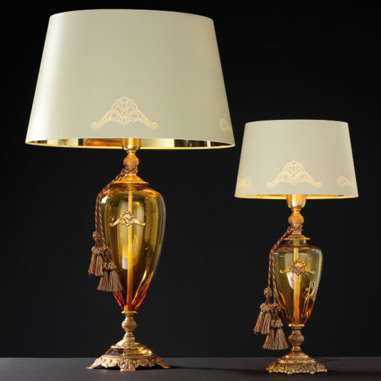 Two Traditional Amber Blown Glass Table Lamps with ornate gold bases stand against a black background. Each elegant lamp features a beige lampshade adorned with delicate gold patterns and tassels, topped with a gold-colored finial. One lamp is larger than the other.