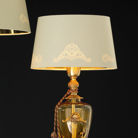 The Traditional Amber Blown Glass Table Lamp features a gold base with intricate, ornate designs set against a black backdrop. It boasts a white lampshade adorned with elegant, symmetrical patterns, while an amber blown glass element enriches its timeless vintage appeal.