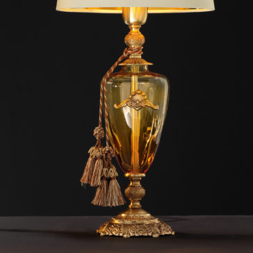 Traditional Amber Blown Glass Table Lamp The Traditional Amber Blown Glass Table Lamp features an ornate design with intricate details and tassels hanging from its neck, all enhanced by a gold-tone finish. Its light-colored lampshade emits a soft glow that beautifully stands out against a dark background.