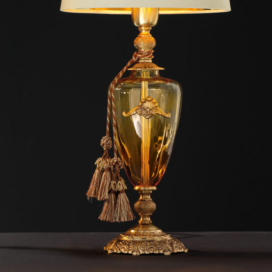 The Traditional Amber Blown Glass Table Lamp features an ornate design with intricate details and tassels hanging from its neck, all enhanced by a gold-tone finish. Its light-colored lampshade emits a soft glow that beautifully stands out against a dark background.