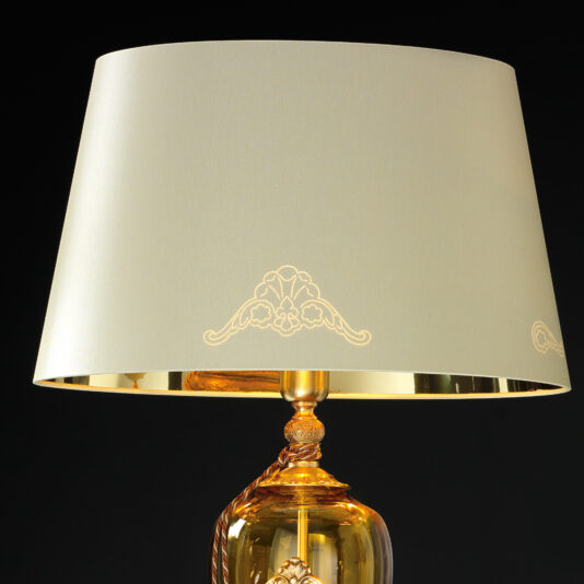 A close-up image highlighting the sophisticated design of the Traditional Amber Blown Glass Table Lamp, featuring a beige lampshade adorned with delicate floral designs. The lamp's base, crafted from transparent amber glass with intricate detailing, stands out against a black background.