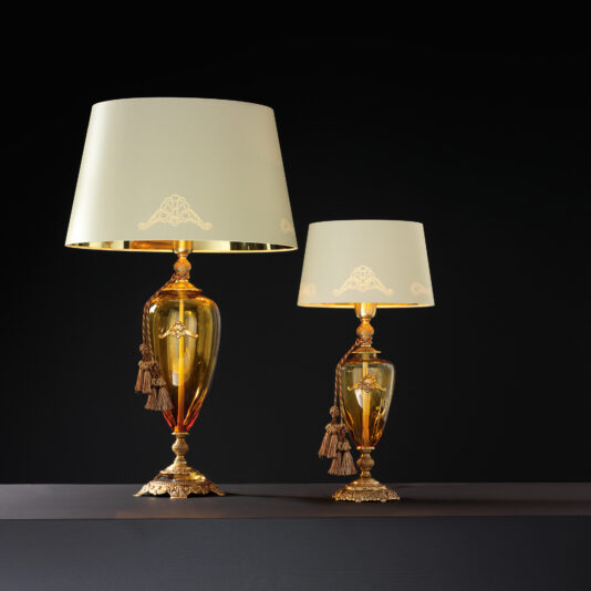 Two elegant, Traditional Amber Blown Glass Table Lamps with beige lampshades and ornate gold bases are displayed against a black background. The larger amber blown glass lamp stands tall beside a smaller, similar lamp. Each base has intricate detailing and tassel decorations, adding a touch of luxury.