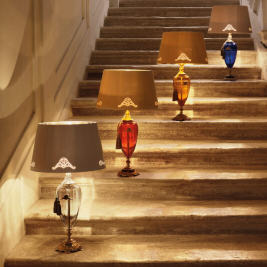 The elegant staircase is illuminated by four decorative lamps, including a Traditional Amber Blown Glass Table Lamp, each with differently colored bases (clear, red, amber, and blue) and beige shades. Positioned on alternating steps, the soft glow of these lamps adds a warm and inviting atmosphere to the scene.