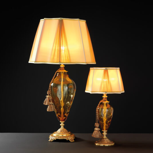 The Traditional Style Dimpled Amber Glass Table Lamps, with their ornate gold bases and dimpled amber glass bodies, exude elegance and tradition. Each lamp features a beige fabric lampshade adorned with decorative tassels. Placed against a dark background, they are illuminated, casting a warm, inviting glow.