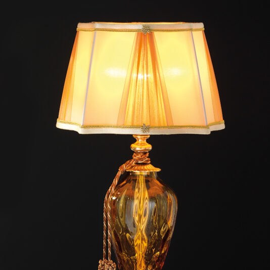Traditional Style Dimpled Amber Glass Table Lamp The Traditional Style Dimpled Amber Glass Table Lamp showcases a golden base with intricate detailing and an ornate lampshade. The gold fabric panels on the shade are adorned with decorative trim, casting a warm, cozy glow when lit.