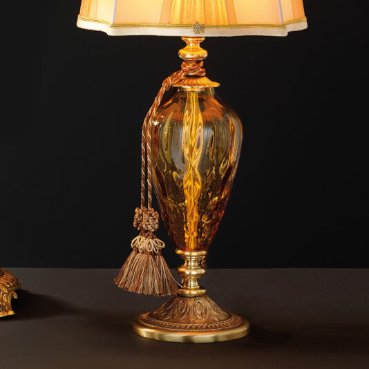 Traditional Style Dimpled Amber Glass Table Lamp The Traditional Style Dimpled Amber Glass Table Lamp boasts an ornate amber glass base with a decorative gold stand and finial. It is crowned with a pleated white lampshade, complemented by a gold tassel hanging from the neck. The black background accentuates its exquisite details.