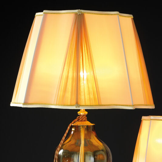Traditional Style Dimpled Amber Glass Table Lamp The Traditional Style Dimpled Amber Glass Table Lamp features a golden base and a large, angular beige lampshade that emits a warm glow. This elegant lamp boasts subtle gold detailing on both the lampshade and base, positioned against a black background for an eye-catching contrast.