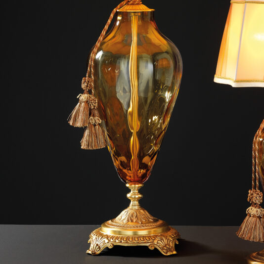 Traditional Style Dimpled Amber Glass Table Lamp The Traditional Style Dimpled Amber Glass Table Lamp, with its ornate gold detailing and decorative tassels, stands elegantly on a black surface against a dark background. The lamp is paired with a cream-colored lampshade trimmed in gold, exuding a vintage and luxurious aesthetic.