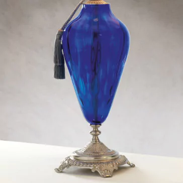 Traditional Style Dimpled Blue Glass Table Lamp The Traditional Style Dimpled Blue Glass Table Lamp is displayed on a white surface, featuring an elegant, elongated shape with a decorative tassel hanging from its neck. The metallic base has intricate detailing and an antique appearance, reminiscent of a traditional-style lamp without the light.