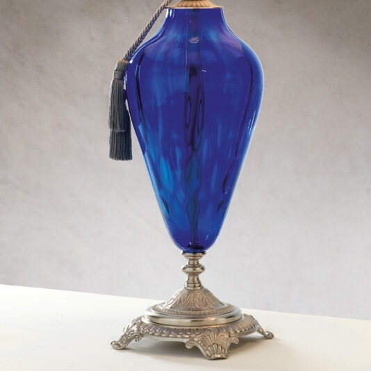 The Traditional Style Dimpled Blue Glass Table Lamp is displayed on a white surface, featuring an elegant, elongated shape with a decorative tassel hanging from its neck. The metallic base has intricate detailing and an antique appearance, reminiscent of a traditional-style lamp without the light.