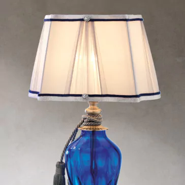 Traditional Style Dimpled Blue Glass Table Lamp The Traditional Style Dimpled Blue Glass Table Lamp features a dimpled blue glass base with a metal accent at the top, wrapped in a rope-like ornament that ends in a tassel. It comes with a beige lampshade adorned with blue trim, which is slightly translucent to allow light to shine through beautifully.