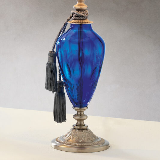 A decorative traditional-style table lamp with a dimpled blue glass body and a metallic base and top, featuring ornate designs. The lamp is adorned with a gray rope tied around the neck, ending in two tassels that hang along its side. Set against a plain, light-colored surface, this elegant piece resembles an intricate vase.
