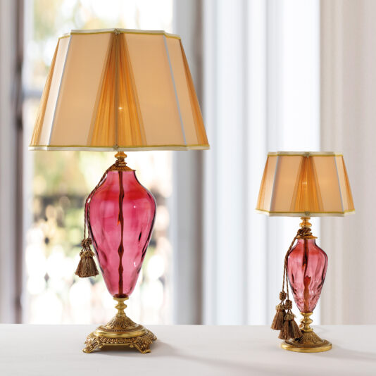 The Traditional Style Dimpled Rose Glass Table Lamps feature two elegant, traditional-style lamps with dimpled rose glass bases and beige, gold-trimmed lampshades. The ornate bases are adorned with gold accents and tassels. Notably, one lamp is significantly larger than the other. Placed on a white surface, they create a harmonious contrast.