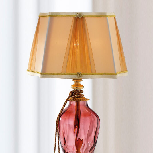The Traditional Style Dimpled Rose Glass Table Lamp features a pink base and a beige pleated lampshade adorned with subtle gold trim. A decorative golden rope wraps around its neck, and in the background, there is a light-colored curtain.