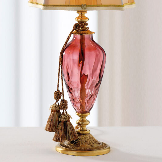 The Traditional Style Dimpled Rose Glass Table Lamp features a red vase-like base adorned with a brown tassel. It stands on an ornate, gold-colored stand and is topped with a beige lampshade trimmed in gold at both ends, all set against a light background.