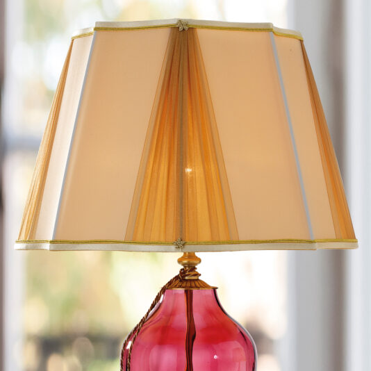 The Traditional Style Dimpled Rose Glass Table Lamp features a gold-trimmed, beige pleated lampshade on a decorative dimpled rose glass base. The softly blurred background hints at an indoor setting with natural light streaming through the windows.