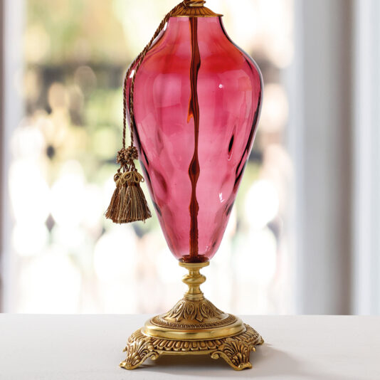 The Traditional Style Dimpled Rose Glass Table Lamp, featuring a crimson dimpled rose glass body and an intricate gold base, stands on a white surface. It is complemented by a braided cord with a tassel. The softly blurred background enhances the lamp's elegant design.
