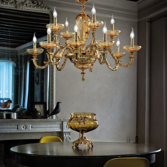 Ornate Jewelled Glass Centrepiece An ornate, gold chandelier with multiple candle-like bulbs hangs above a round dining table adorned with an Ornate Jewelled Glass Centrepiece. The table also features a gold, intricate vase. Behind the table, there is a fireplace with various decorative items and a large mirror. A black bird statue rests on the mantel.
