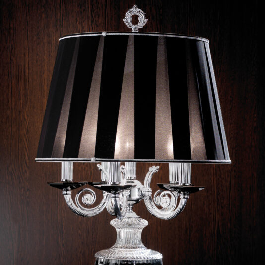 Candelabra Style Large Glass Table Lamp The Candelabra Style Large Glass Table Lamp boasts a stylish large glass build along with a black and silver striped lampshade. Its ornate base features an intricate glass design and multiple candle-like holders beneath the shade, set against a dark wood-paneled wall.