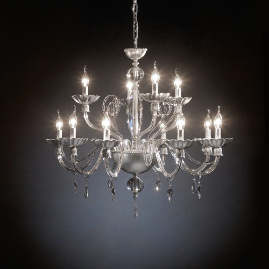 Candle Style Chandelier With Crystal Drops The Candle Style Chandelier With Crystal Drops, a sophisticated light fixture with twelve elegantly curved arms each featuring a candle-shaped light bulb, hangs against a dark backdrop. Its intricate design glistens beautifully as it fills the space with a warm, ambient glow.