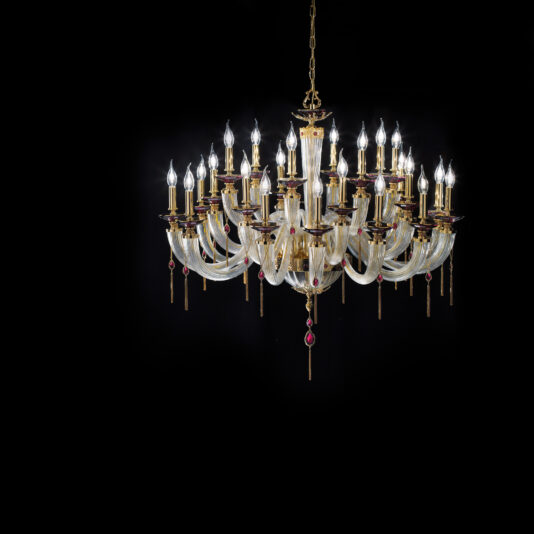 The Candle Style Chandelier With Swarovski® Crystals is a large, elegant fixture that extends from a gold frame and features multiple tiers of candle-like bulbs. It boasts curved glass arms and red Swarovski® crystal accents, all set against a black background.