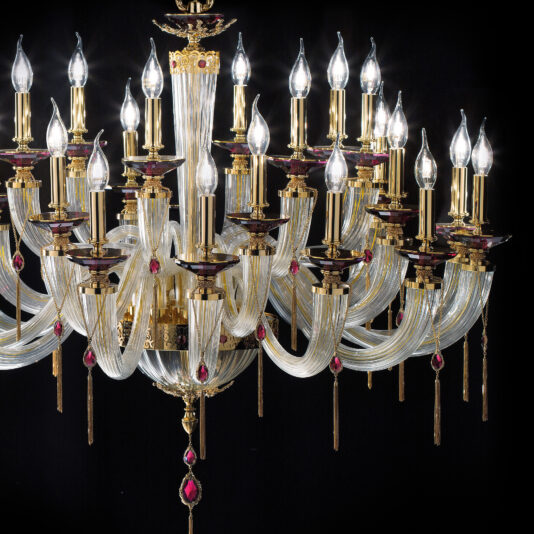 The Candle Style Chandelier With Swarovski® Crystals boasts multiple gold arms and candle-like bulbs, each adorned with red gemstone accents. Its intricate design includes twisted Swarovski® crystal strands and hanging decorative elements, offering a luxurious and opulent appearance against a black background.