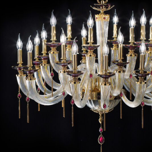 The Candle Style Chandelier With Swarovski® Crystals is a luxurious lighting fixture featuring numerous candle-like light bulbs arranged in ornate golden holders. This chandelier is adorned with intricate glass arms and decorative red gemstones, enhanced with dazzling Swarovski® crystals, all set against a dark background. The bulbs emit a soft, elegant glow.