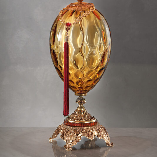 Classic Amber Dimpled Glass Table Lamp An elegant Classic Amber Dimpled Glass Table Lamp sits atop an ornate metal base with intricate filigree details. A long red tassel with a decorative chain drapes over the top, adding a touch of luxury and sophistication. The background is a neutral gray.