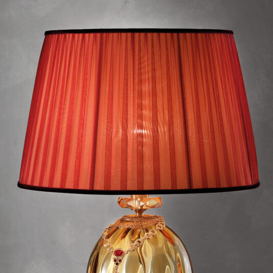 Classic Amber Dimpled Glass Table Lamp Presenting the Classic Amber Dimpled Glass Table Lamp, a golden table lamp with an ornate base adorned with red and gold accents and featuring a ruby-like gem encased in an intricate design. It boasts a rich red pleated shade that offers soft illumination against a gray background.