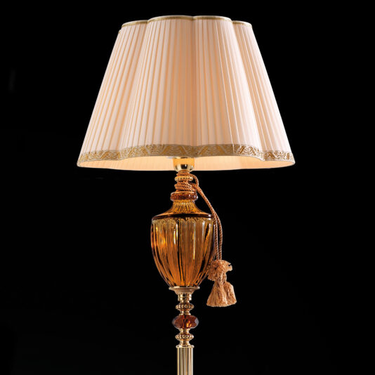 An elegant table lamp adorned with a pleated beige shade, decorative golden base, intricate detailing, and a hanging tassel captures the sophisticated style of the Classic Amber Glass Floor Lamp, showcased against a black background.