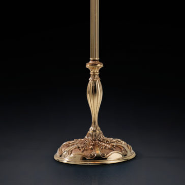 A gold-colored, intricately designed base of a Classic Amber Glass Floor Lamp is centered against a dark background. The ornate details include a ribbed column and an elaborate, decorative circular base with floral and curved patterns.