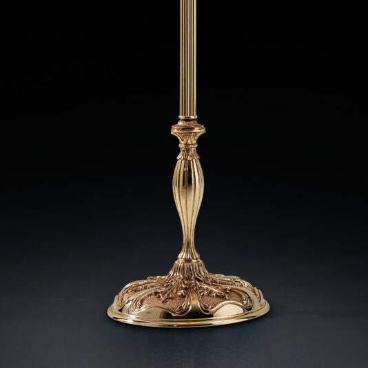 A gold-colored, intricately designed base of a Classic Amber Glass Floor Lamp is centered against a dark background. The ornate details include a ribbed column and an elaborate, decorative circular base with floral and curved patterns.