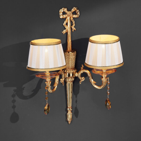 The Classic Style Wall Light With Swarovski® Crystals is a gold, ornate two-light wall sconce adorned with cream-colored lampshades and detailed with hanging Swarovski® crystals. Mounted on a dark gray wall, its design showcases intricate scrollwork and a decorative central element featuring vertical ridges.