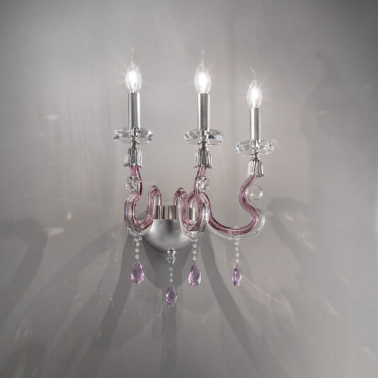 The Contemporary 3 Arm Candle Style Glass Wall Light is a decorative fixture that showcases sophisticated curves in pink and clear glass, adorned with hanging pink crystal pendants. This wall-mounted light features three candle-shaped bulb holders, casting delicate shadows against a plain gray backdrop.